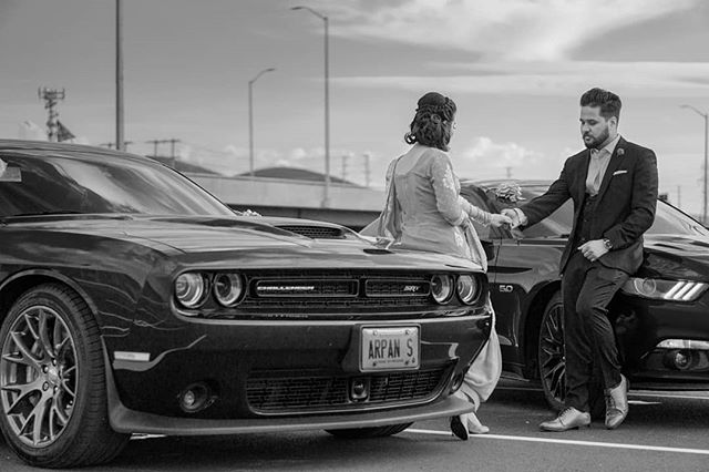 A couple poses with a Dodge Challenger and a Ford Mustang in a black and white shot.