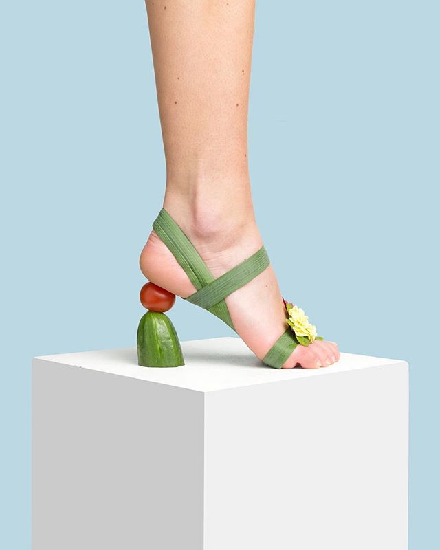 A woman's foot wears a sandal made of green leaves, a tomato, and cucumber, creatively displayed on a white cube.