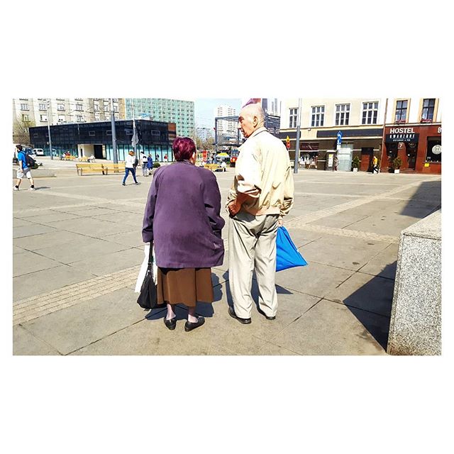 An elderly couple stands in a city square on a sunny day, enjoying retirement.