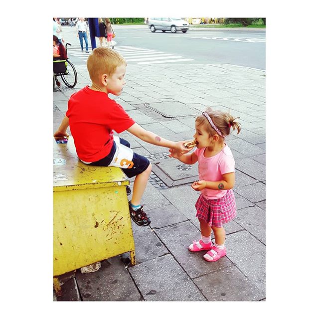 A young boy shares a donut with a girl on a city sidewalk, in a candid and cheerful moment.