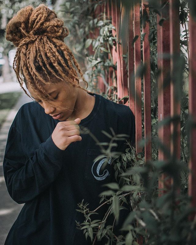 A young woman with dreadlocks tied up in a bun stands pensively by a fence.