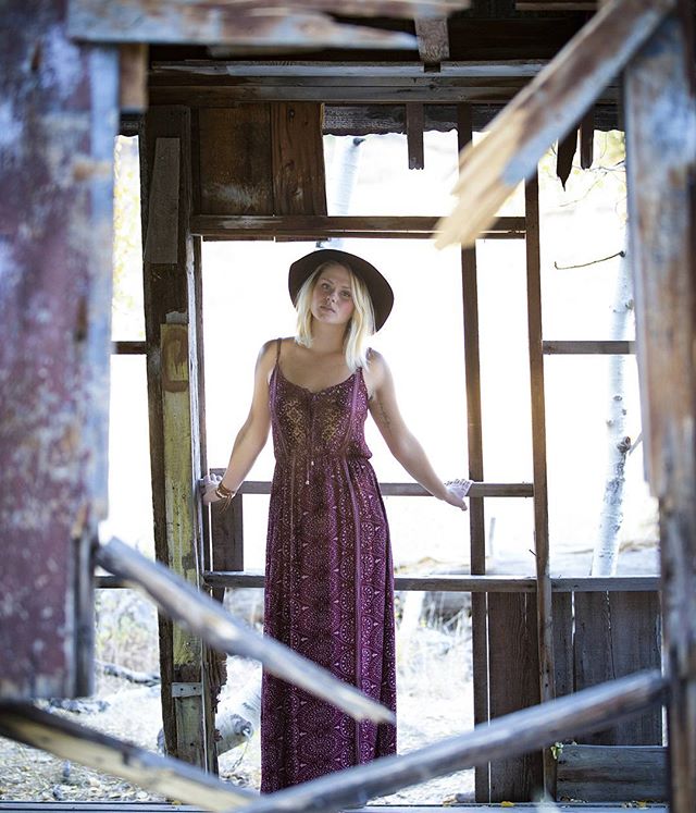 A woman wearing a dress and hat poses in the frame of an abandoned building.