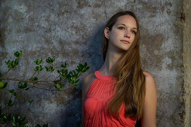 A serene woman in a red dress poses against a textured wall in a thoughtful, outdoor portrait.