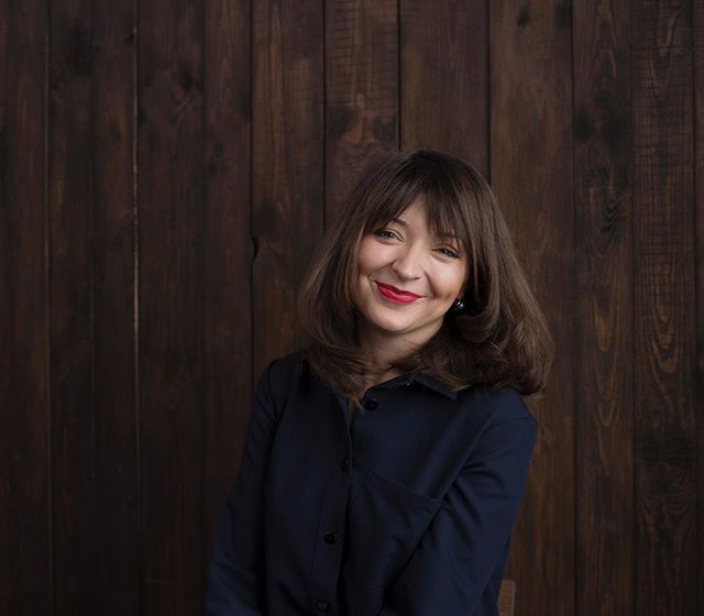 A smiling brunette woman poses against a wooden background, projecting a relaxed and approachable persona.