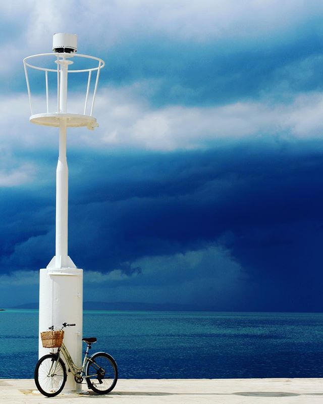 A bicycle stands near a white lamp post by the ocean under a dramatic, cloudy sky.