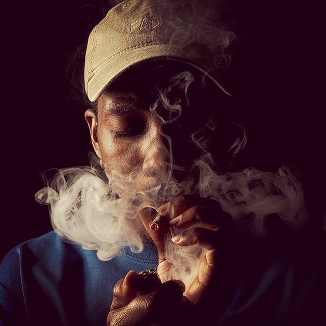 A man in a cap exhales smoke from a cigarette in a darkly lit indoor setting.