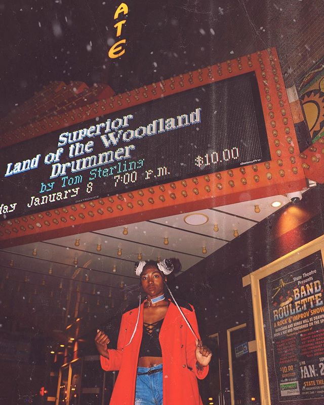 A woman stands under a vintage theater marquee promoting a local show at night.