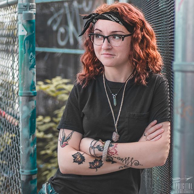 A young woman with red hair and tattoos poses confidently against a chain-link fence.