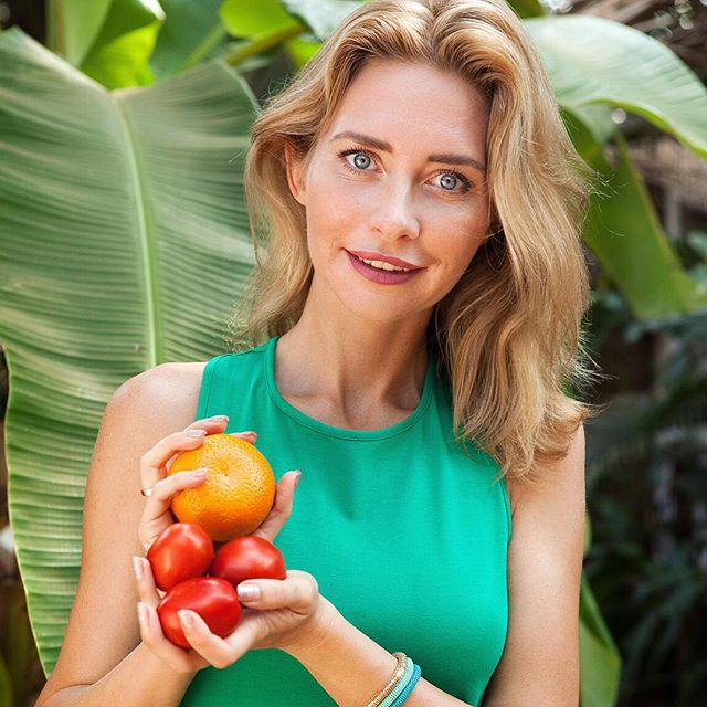 A woman holds an orange and tomatoes outdoors surrounded by lush greenery.