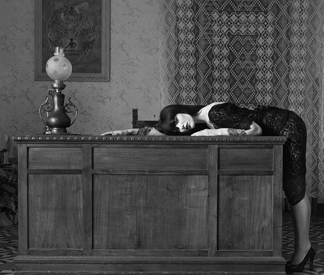 A woman in a lace dress poses on a vintage desk with a lamp in a moody, black and white editorial style.
