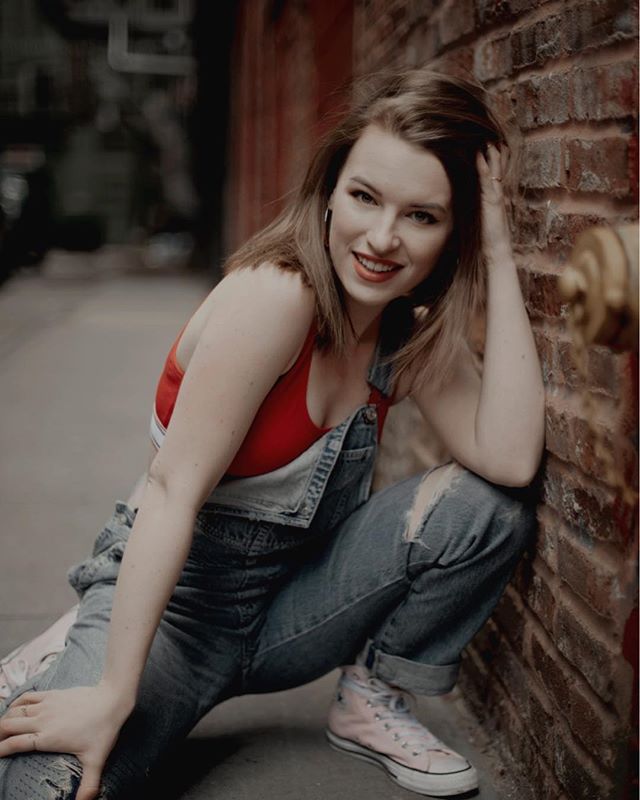 A smiling woman in overalls and a red top poses against a brick wall in an urban alleyway.