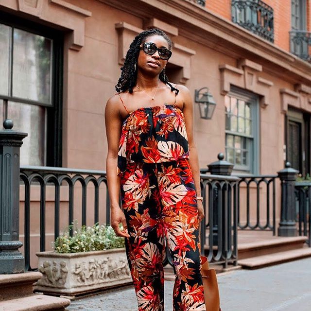 A stylish woman in a floral jumpsuit and sunglasses walks down a city street.