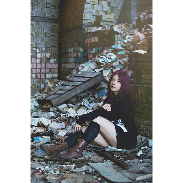 A woman with purple hair sits among scattered books in an abandoned building, creating a moody editorial scene.