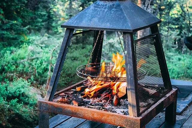 A metal fire pit with flames and firewood burns outdoors surrounded by greenery. 