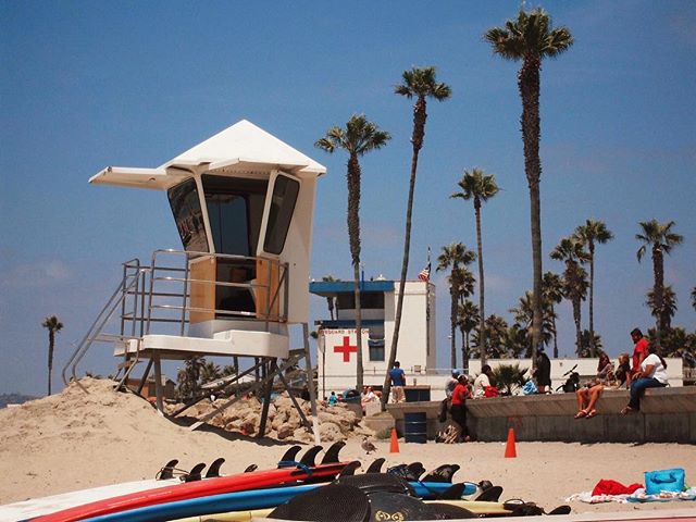 A sunny beach scene with a lifeguard tower, palm trees, and people relaxing near surfboards.