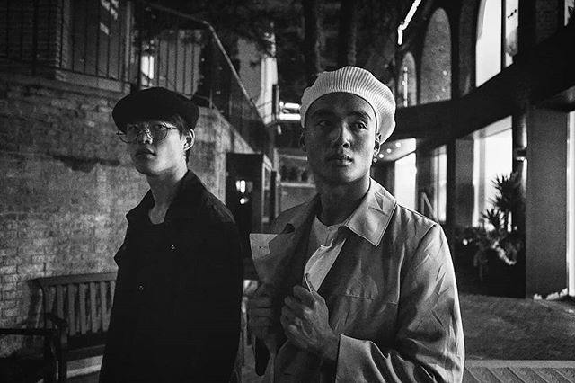 Two men pose outdoors in black and white, wearing hats and coats in an urban setting at night.