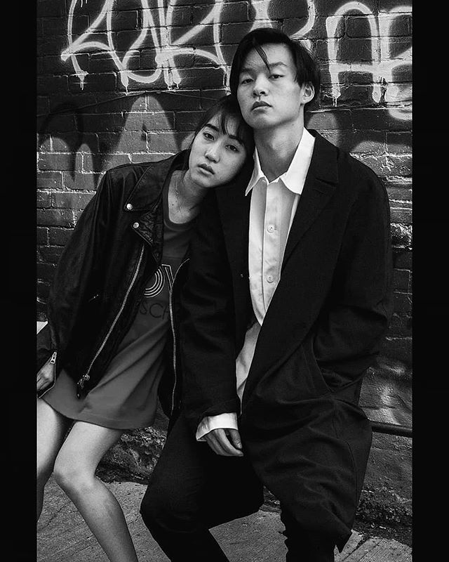 A stylish couple poses against a graffiti-covered brick wall in a black and white editorial style.