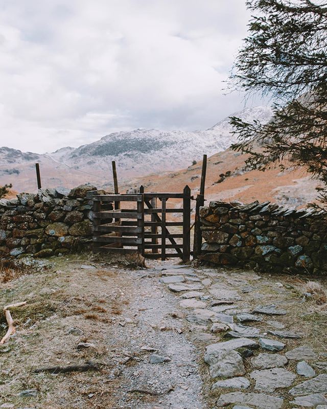 A stone pathway leads through a wooden gate in a stone wall, with mountains visible in the distance under a cloudy sky.