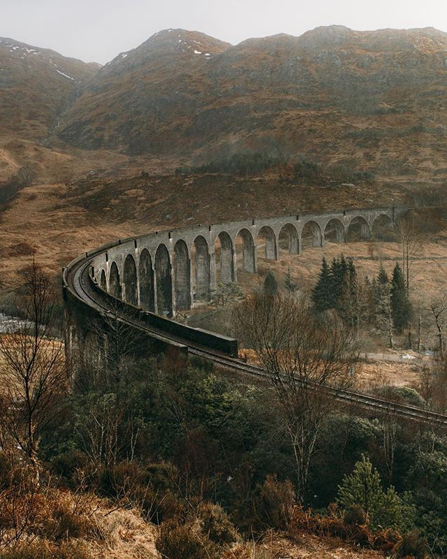 The Glenfinnan Viaduct, a historic railroad bridge, spans a scenic Scottish valley with mountains in the background.