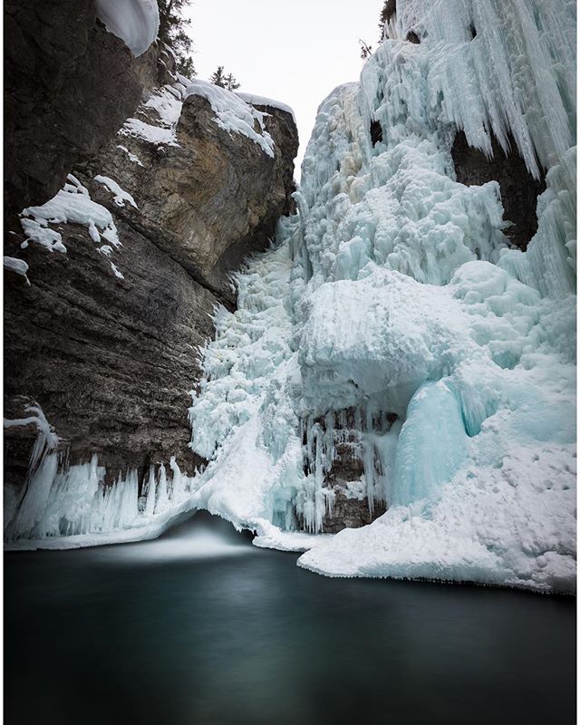 A frozen waterfall cascades into a dark pool in a dramatic, snowy canyon.