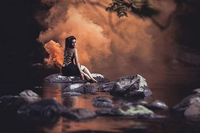A woman in a dress sits on rocks in the water, surrounded by smoke in a dreamy, outdoor scene.