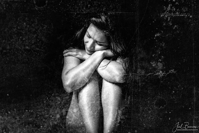 A woman sits hugging her legs in a moody, intimate black and white studio portrait.
