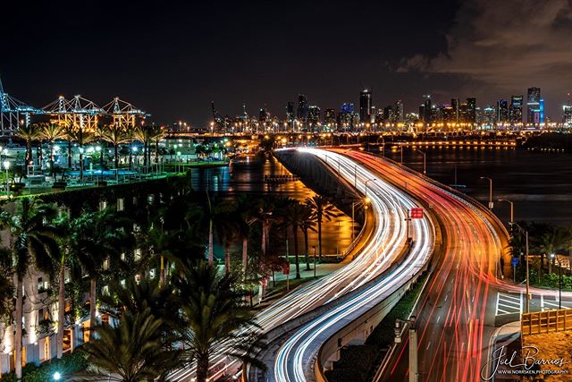 Miami skyline at night with long exposure traffic on a bridge, showcasing vibrant city lights and waterfront views.