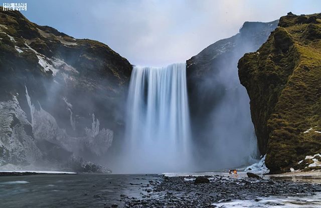 A scenic view of a powerful waterfall cascading through mountains in Iceland with a dog exploring the stony beach.
