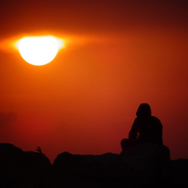 A person's silhouette sits serenely against a vibrant orange sunset during outdoor meditation.