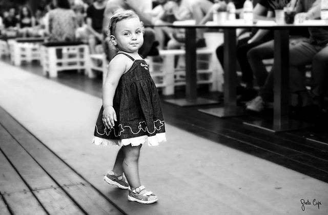 A young girl in a dress walks confidently in a black and white image, surrounded by people enjoying a restaurant.