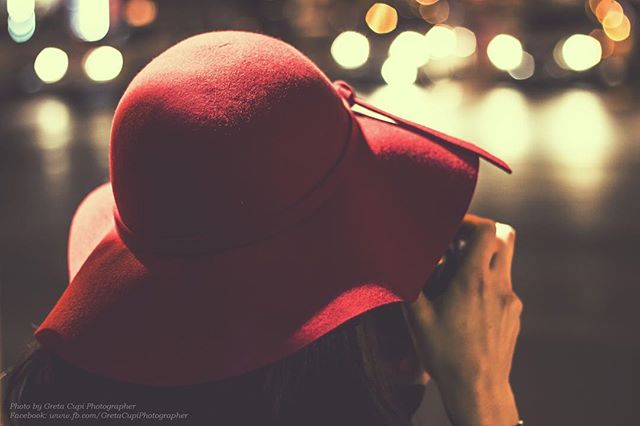 A woman in a red hat takes pictures at night in the city with blurred city lights in the background.