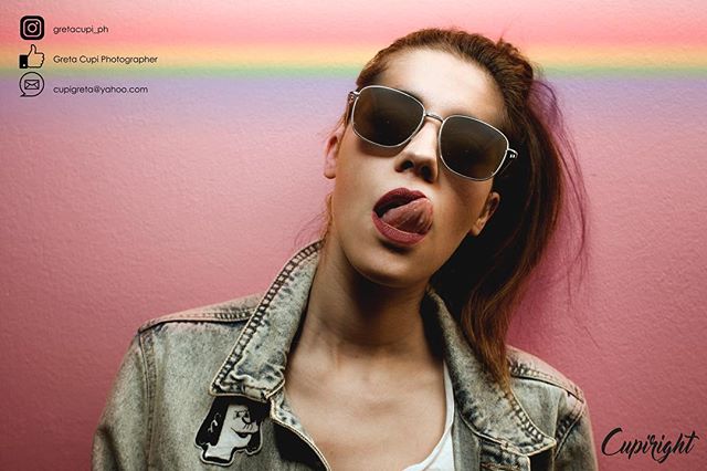 A stylish woman poses with sunglasses and a denim jacket against a pink backdrop with a playful expression.
