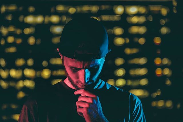 Contemplative man in a cap stands against a bokeh cityscape, lit with neon hues in an urban setting.