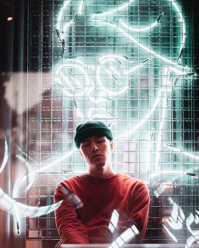 A man wearing a beanie and sweater poses in front of a glowing neon sign at night.