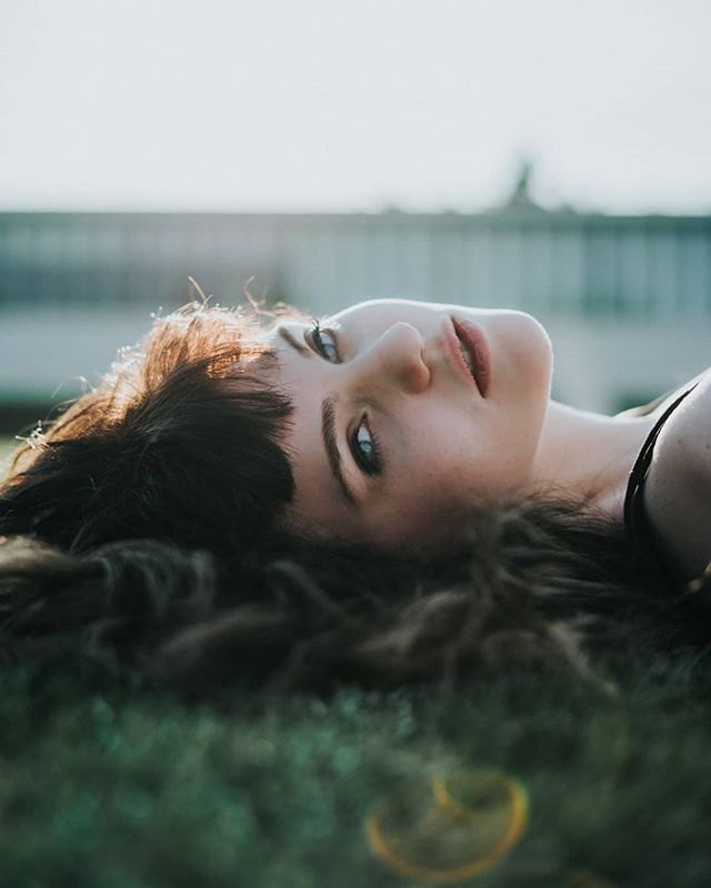 A brunette woman with bangs lies on the grass, looking up with a dreamy and relaxed expression.
