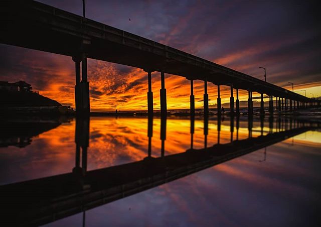A tranquil sunset reflected in the water under a silhouetted pier, capturing the beauty of a beach scene.