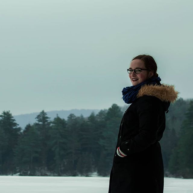 A woman in a black coat and blue scarf smiles on a snowy landscape with a forest in the background.
