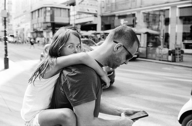 A father carries his daughter piggyback down a city street, in a candid black and white moment.