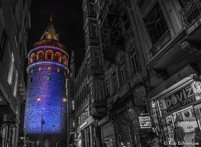 The Galata Tower is illuminated at night on a narrow city street in Istanbul, Turkey.