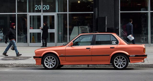 An orange BMW E30 parked on an urban street with people walking by, showcasing classic car culture.