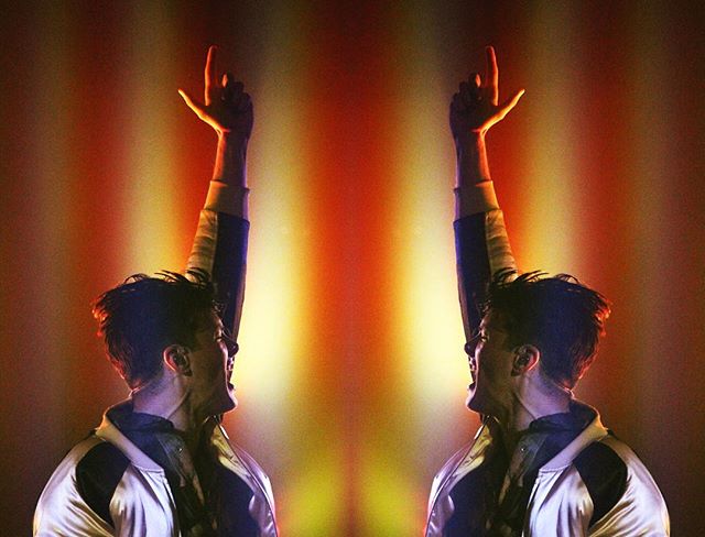 A man with arms raised performs on stage at a concert with bright, vibrant lights.