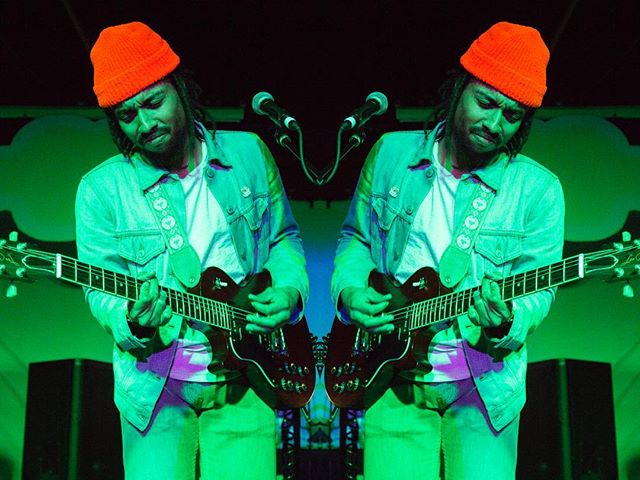 Musician playing guitar on stage under green lights, wearing an orange hat, mirrored.