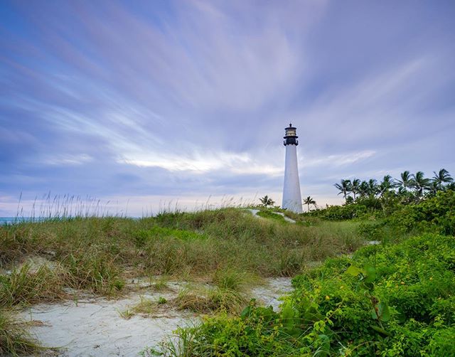 A scenic view of a lighthouse on a sandy beach with grassy dunes, beneath a cloudy sky at dusk or dawn.