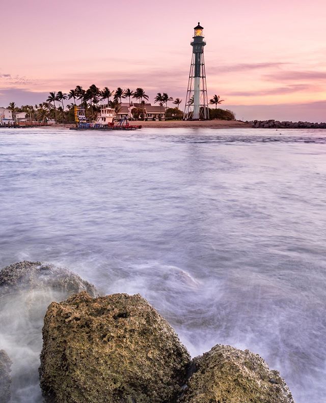 A serene view of a lighthouse and a boat near palm trees during a colorful sunset at the beach.