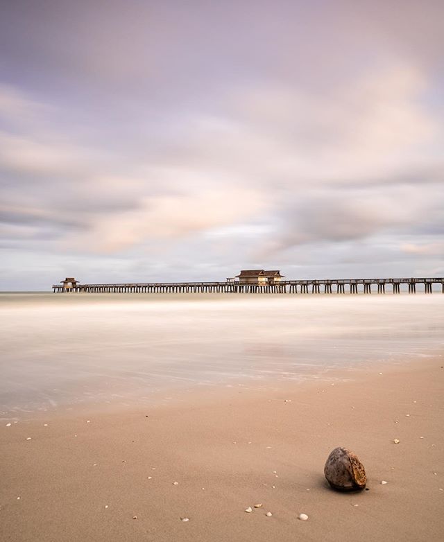 A scenic view of a pier extending into the ocean with a coconut on the sandy beach under a cloudy sky.