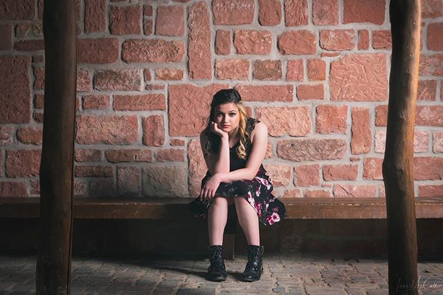 A woman in a dress sits thoughtfully on a bench against a textured brick wall backdrop.
