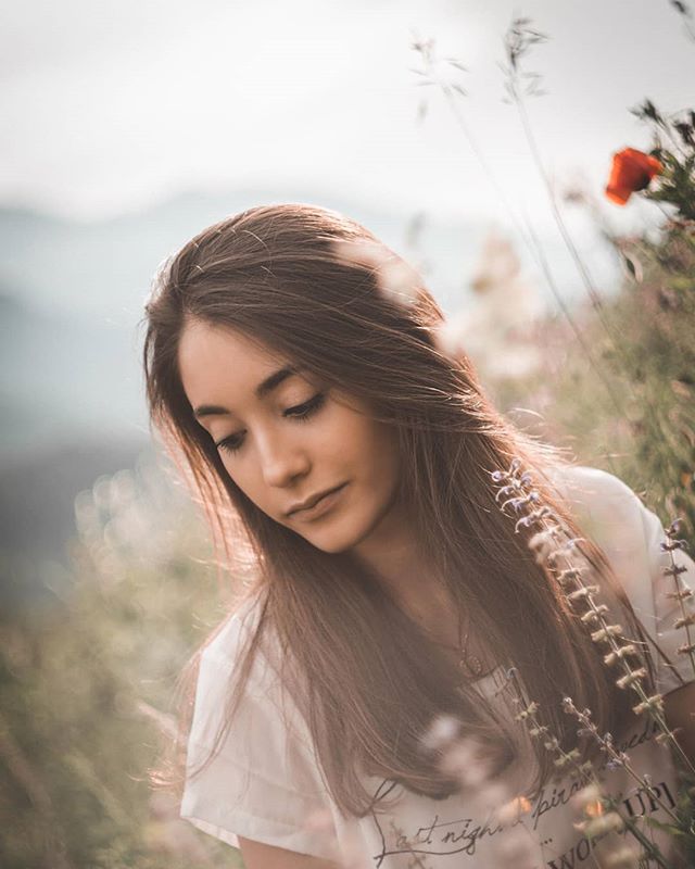 A young woman with brown hair looks down in a field of flowers in soft, natural light.