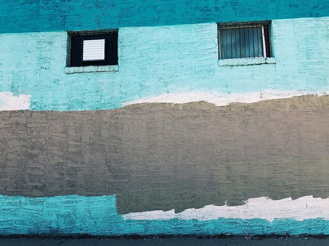 A textured building wall featuring blue paint, a gray section, and two windows.