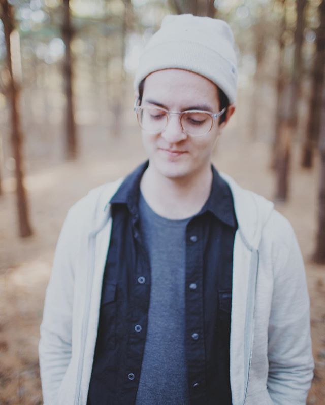 A person with glasses and a beanie looks down in a wooded area, a soft light illuminating the scene.