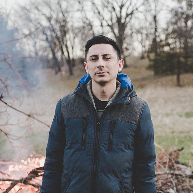 A man in a blue jacket stands outdoors with trees in the background, exuding a calm and contemplative mood.